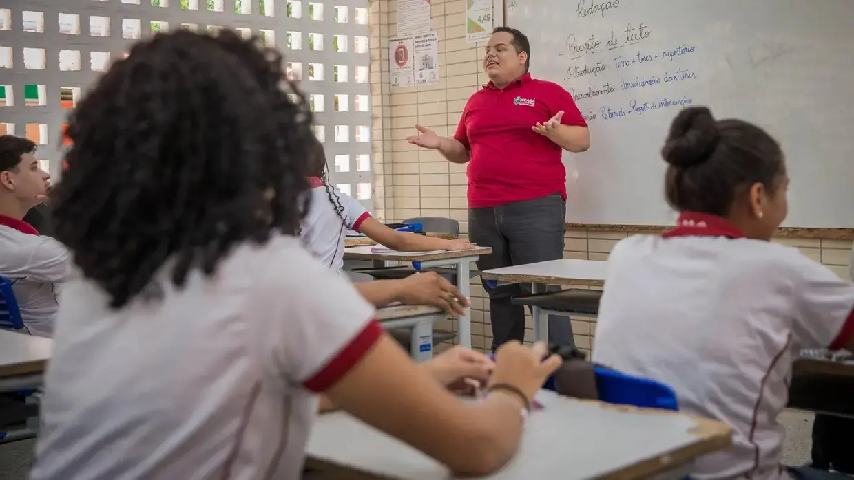 professor em sala de aula, diante de professores. atrás dele, uma tela com textos escritos.