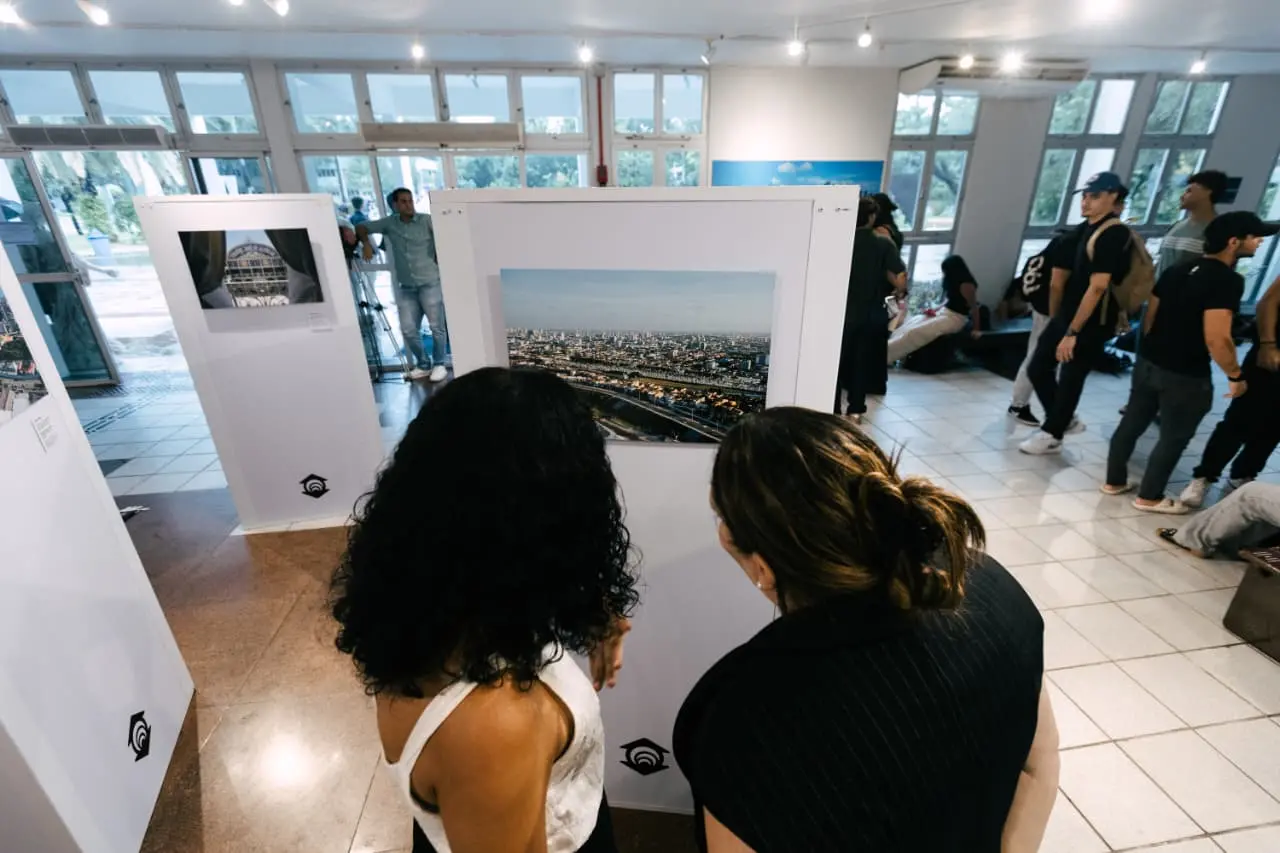 Na imagem, uma fotografia de plano médio, tirada de um ângulo superior, mostra duas mulheres de costas observando uma fotografia em uma exposição de arte. A mulher à esquerda tem cabelos pretos e cacheados e veste uma regata branca; a mulher à direita tem o cabelo castanho preso em um coque e veste uma blusa preta com risca de giz. Elas olham para uma fotografia horizontal de uma paisagem urbana aérea, montada em um painel branco vertical. No centro do painel, abaixo da foto, há um logotipo preto pequeno. Ao redor, o ambiente da galeria é amplo, com piso de azulejos claros e iluminação de trilhos no teto. Ao fundo, outras pessoas circulam pela exposição, e grandes janelas de vidro mostram uma área externa arborizada e clara. O clima é de um evento cultural movimentado.