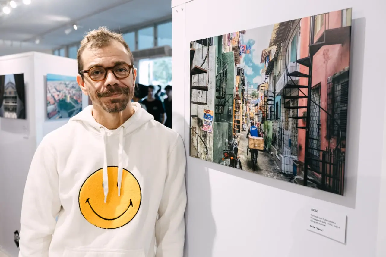 Na imagem, fotografia de plano médio em ambiente de galeria. À esquerda, um homem de pele clara, óculos de armação tartaruga, barba curta e cabelos castanhos levemente grisalhos, olha para a câmera com um sorriso discreto. Ele veste um moletom branco com capuz, que traz no centro uma grande estampa amarela do ícone 
