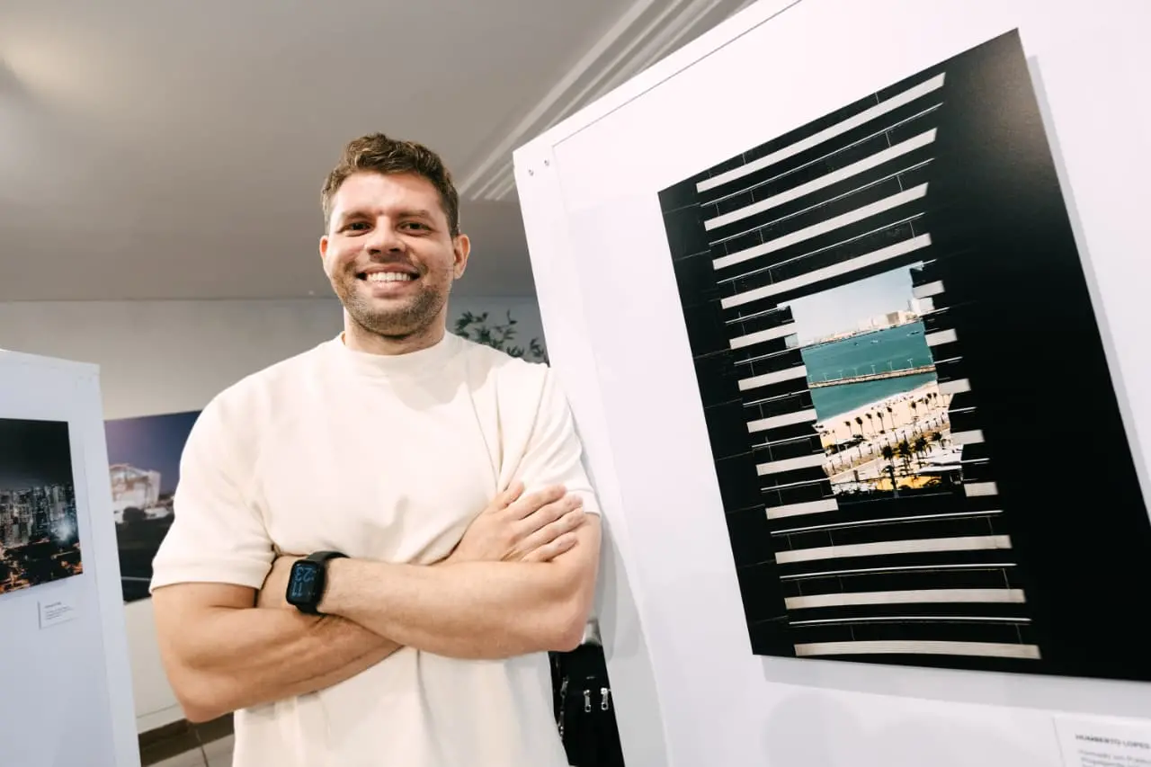 Na imagem, fotografia de plano médio em ângulo levemente de baixo para cima. À esquerda, um homem de pele clara, cabelos curtos castanhos e barba rala sorri para a câmera com os braços cruzados. Ele veste uma camiseta branca de gola redonda e usa um relógio digital preto no pulso esquerdo. À direita, em destaque, há uma obra fotográfica montada em um painel branco. A fotografia é vertical e possui uma moldura ou grafismo em preto com linhas horizontais vazadas, criando o efeito de 