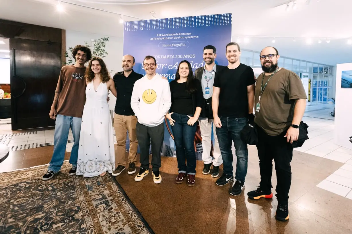 Na imagem, uma fotografia de plano médio mostra um grupo de oito pessoas — seis homens e duas mulheres — posando sorridentes em um ambiente de galeria de arte. Eles estão dispostos lado a lado à frente de um painel azul vibrante que contém textos institucionais sobre a 