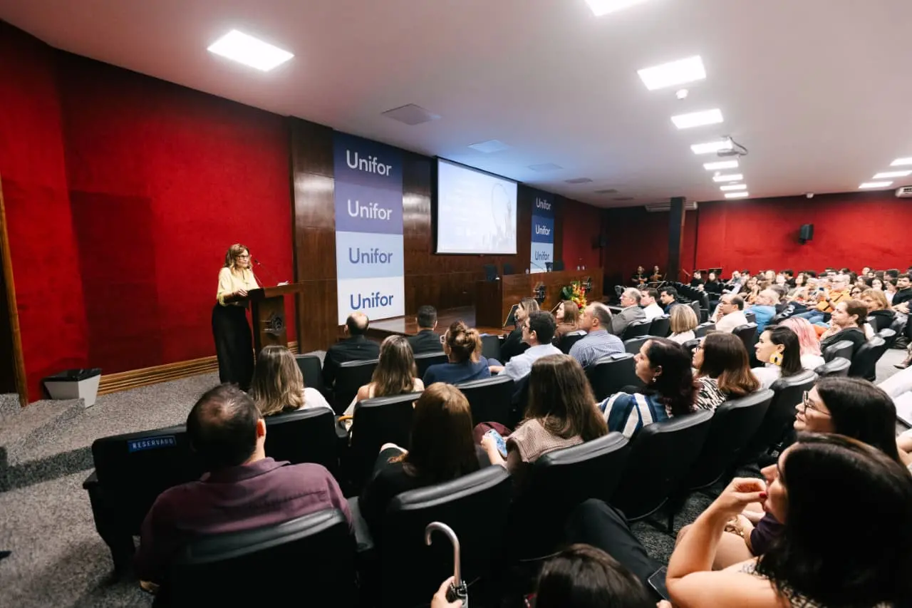 Na imagem, fotografia em plano aberto de um auditório lotado durante um evento acadêmico ou institucional. A imagem é capturada de uma perspectiva elevada, mostrando as fileiras de poltronas pretas ocupadas por uma plateia diversificada, vista majoritariamente de costas. À esquerda, no palco, uma mulher de cabelos loiros e óculos, vestindo uma blusa amarela e calça preta, fala ao microfone em um púlpito de madeira. A parede atrás dela é revestida por uma textura vermelha vibrante. Ao centro do palco, há painéis verticais azuis com o logotipo 