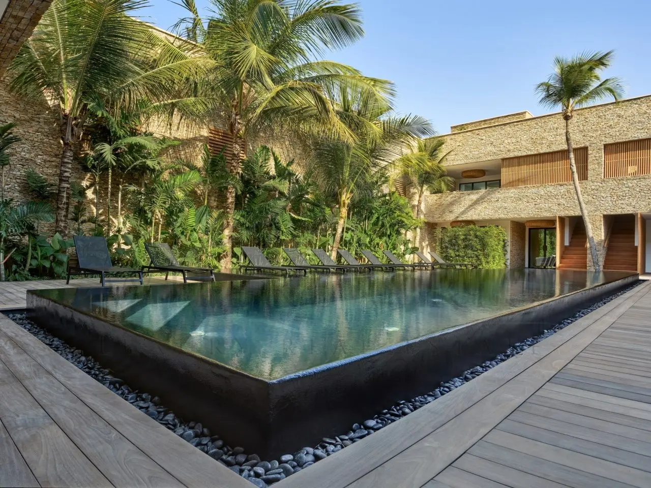 Piscina retangular cercada por espreguiçadeiras e coqueiros, em área externa de casa de luxo com fachada de pedra e madeira.