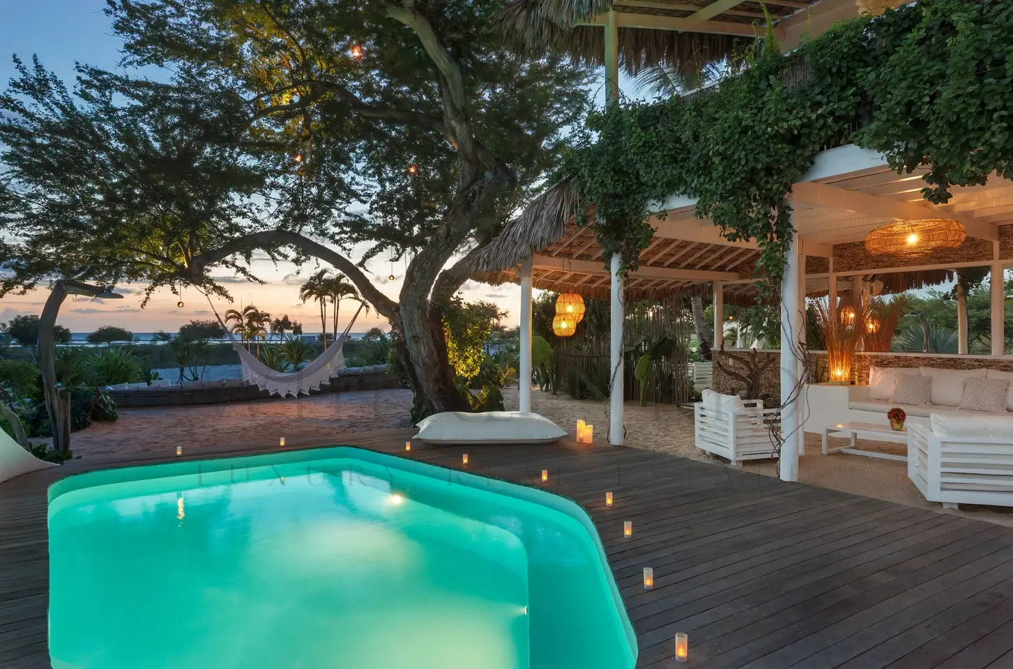 Área externa de casa de praia ao entardecer, com piscina iluminada, varanda com sofás, rede entre árvores e vista para o mar.