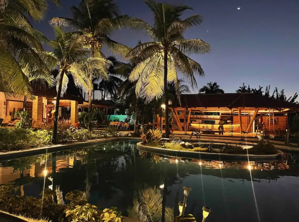 Área externa de casa de praia com árvores, piscina iluminada, varanda e bar.