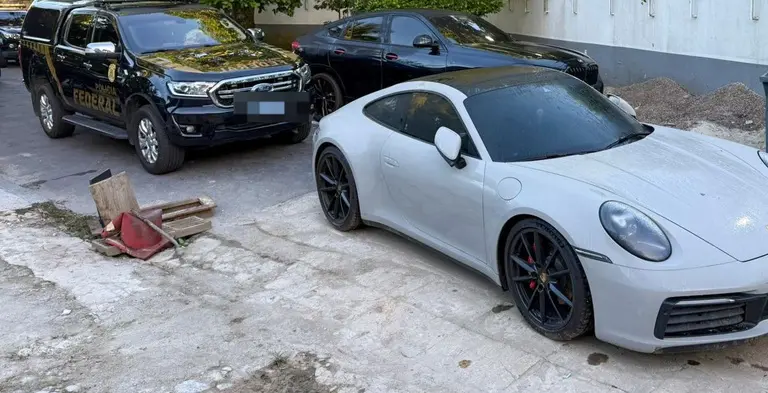Carro esportivo Porsche branco estacionado ao lado de veículos de polícia federal em uma área de estacionamento ao ar livre. A imagem mostra detalhes do ambiente, incluindo alguns objetos no chão e árvores ao fundo.