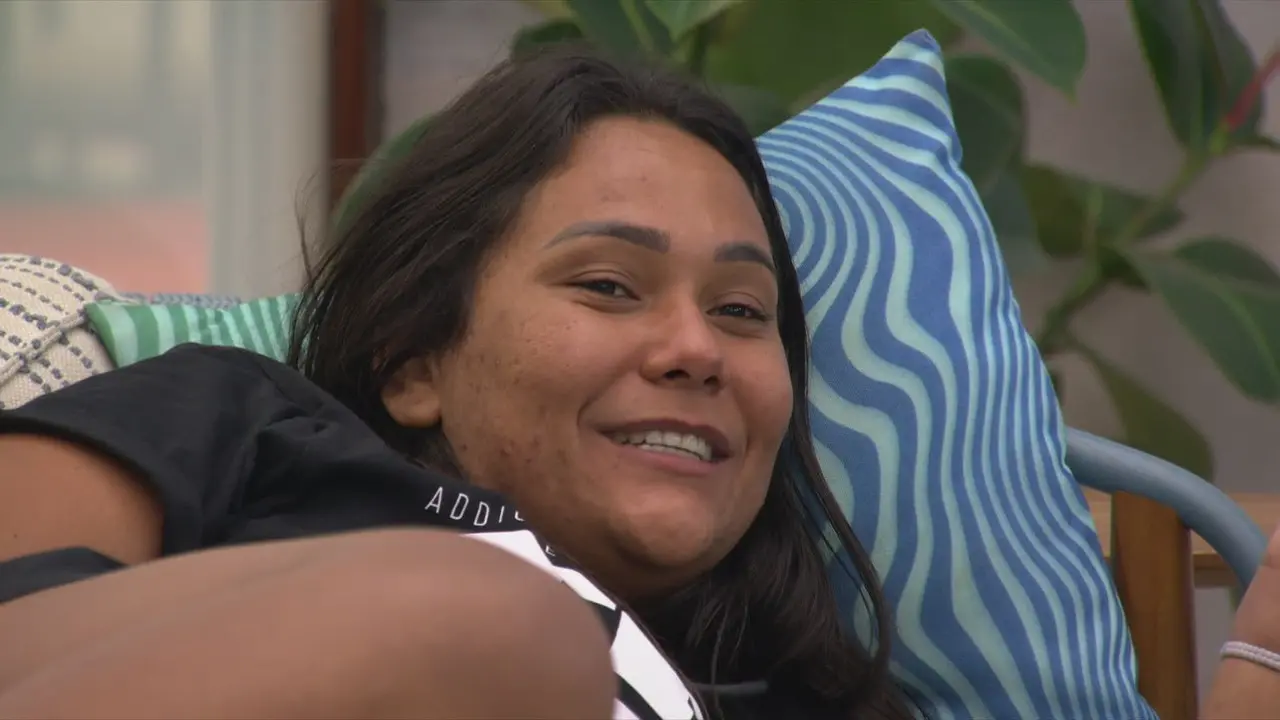 Pessoa deitada em sofá com almofadas estampadas, em área interna, posicionada de lado e olhando em direção à câmera.