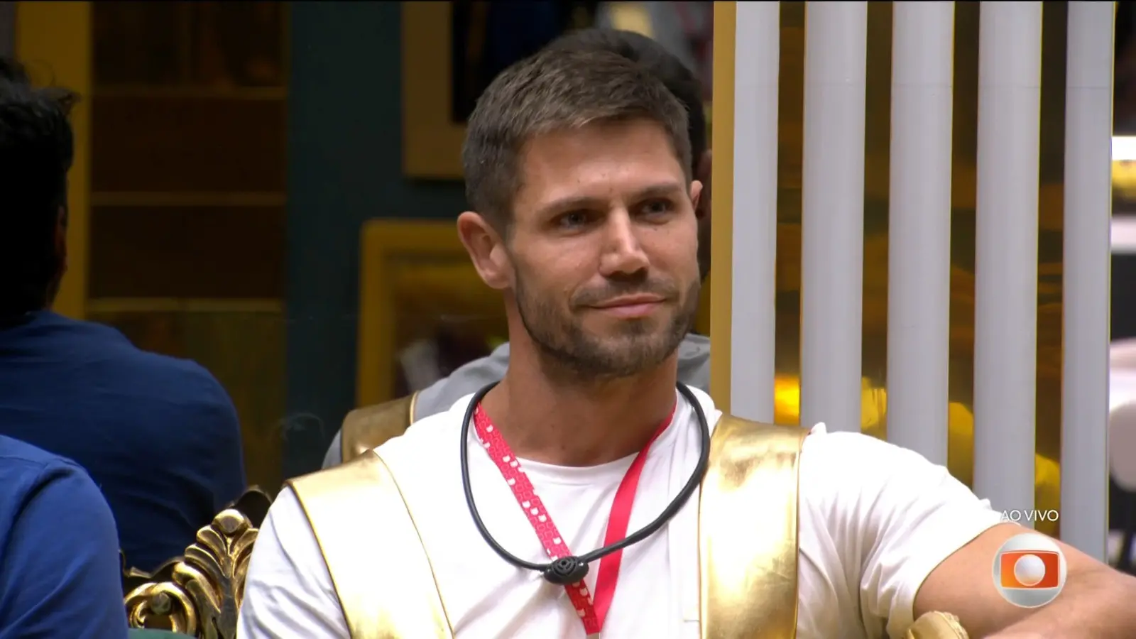 Pessoa sentada em área interna observa ao redor, usando camiseta clara e cordão no pescoço; ao fundo há divisórias verticais e outras pessoas desfocadas.