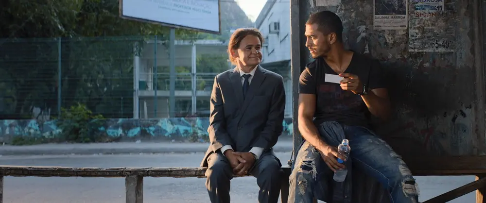 O ator Gero Camilo, vestindo um terno cinza, senta-se ao lado de um jovem, o ator Ruan Aguiar, em um ponto de ônibus desgastado, enquanto ambos trocam olhares e o rapaz segura um pequeno cartão branco. A luz do entardecer ilumina a cena do filme 'Papagaios', destacando o contraste entre o traje formal de um e o estilo casual de calça jeans rasgada do outro.