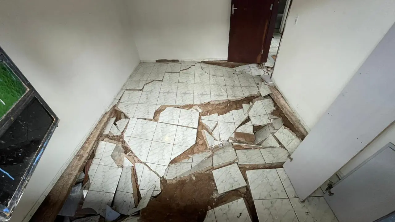 O piso de cerâmica clara de um cômodo está completamente destruído e afundado, revelando a terra e o solo sob a estrutura da casa. Os azulejos quebrados em grandes pedaços se espalham pelo ambiente vazio, evidenciando o grave colapso estrutural próximo a uma porta de madeira e paredes brancas.