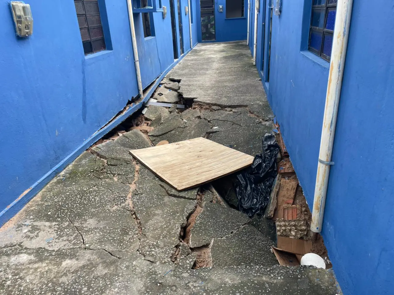 O corredor de cimento entre duas fileiras de casas azuis está gravemente danificado, com grandes rachaduras e buracos que revelam o solo por baixo. Madeira e sacos plásticos pretos cobrem parcialmente uma fenda no chão danificado. As paredes azuis das casas, com janelas e portas visíveis, dão para o corredor. Um tubo de escoamento branco sobe pela parede direita. O corredor se estende para longe, revelando mais danos ao cimento e o final da rua.