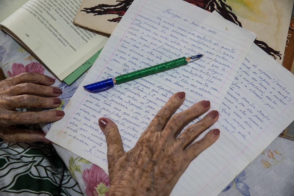 Na imagem, vista superior em close-up das mãos de uma pessoa idosa sobre uma folha de papel pautado com um texto escrito à mão em tinta azul. As mãos têm unhas pintadas de esmalte rosado e a pele apresenta marcas do tempo. Sobre o papel, repousa uma caneta de corpo verde brilhante com tampa azul. Ao lado, vê-se parte de um livro aberto e uma toalha de mesa com estampa floral. O título do texto manuscrito é