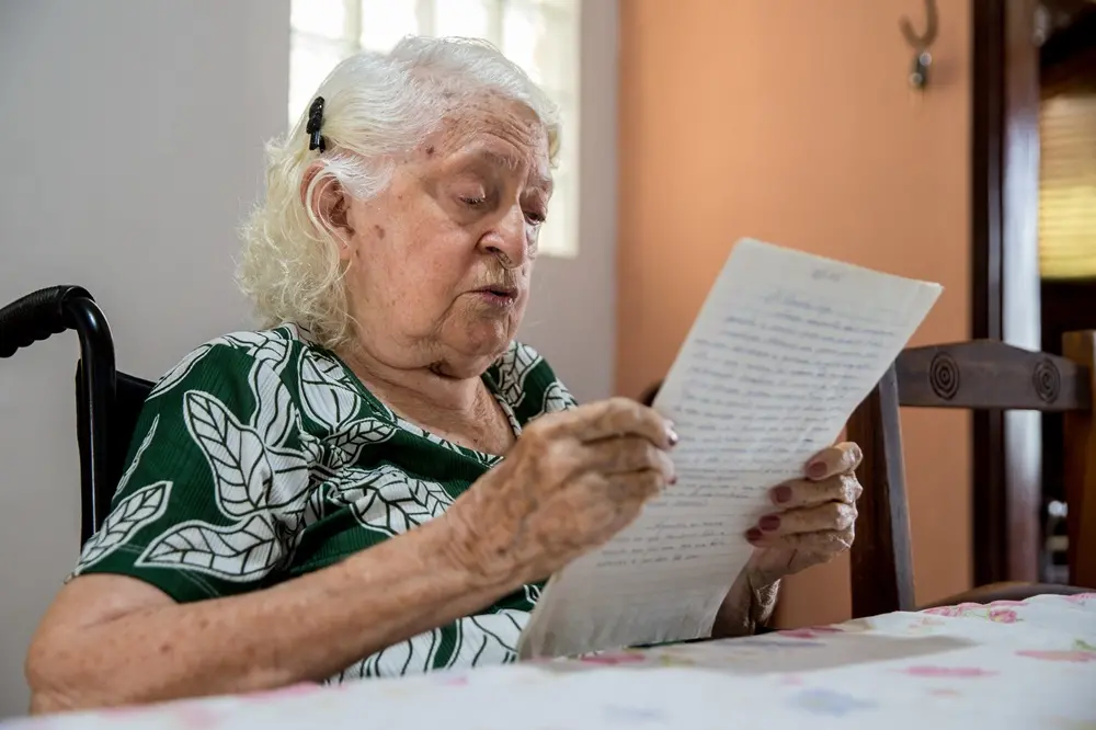 Na imagem, uma mulher idosa de cabelos brancos, sentada em uma cadeira de rodas, segura e lê atentamente uma folha de papel pautado com escrita manuscrita. Ela veste uma blusa verde com estampa de folhagens brancas. A cena acontece em uma sala iluminada; ao fundo, vê-se uma parede de tom pêssego e parte de uma cadeira de madeira. A mulher está de perfil parcial, concentrada na leitura do documento que segura com as duas mãos sobre uma mesa com toalha florida.
