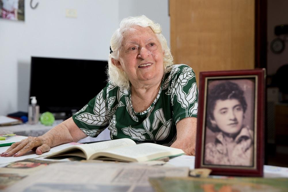 Na imagem, uma mulher idosa de cabelos brancos e blusa verde estampada sorri enquanto olha para algo fora do campo de visão. Ela está sentada à mesa com um livro aberto e papéis à sua frente. No primeiro plano, à direita, há um porta-retrato com uma fotografia antiga em preto e branco de uma mulher jovem. O fundo mostra o interior de uma casa com uma televisão e uma porta de madeira. A iluminação é suave e frontal.