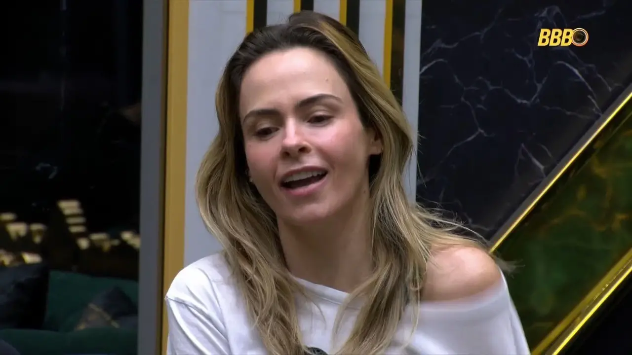 Pessoa sentada em ambiente interno do programa BBB, usando blusa branca, vista em plano médio; ao fundo, paredes com painéis geométricos escuros e o logotipo “BBB” no canto superior direito.