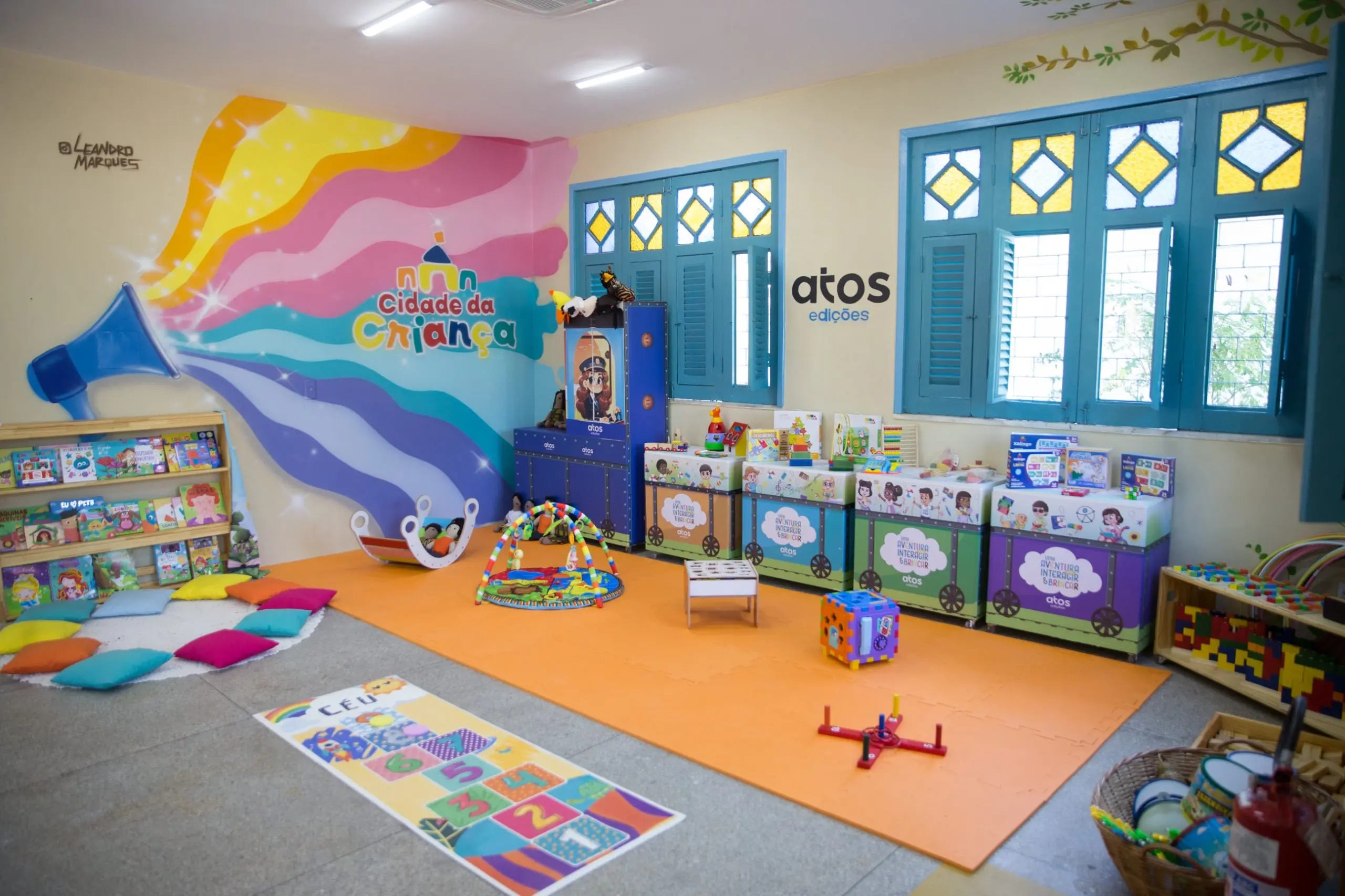 Brinquedoteca colorida com tapete laranja, estante de livros, amarelinha no chão e móveis temáticos. A parede exibe uma pintura de arco-íris e o logotipo Cidade da Criança.