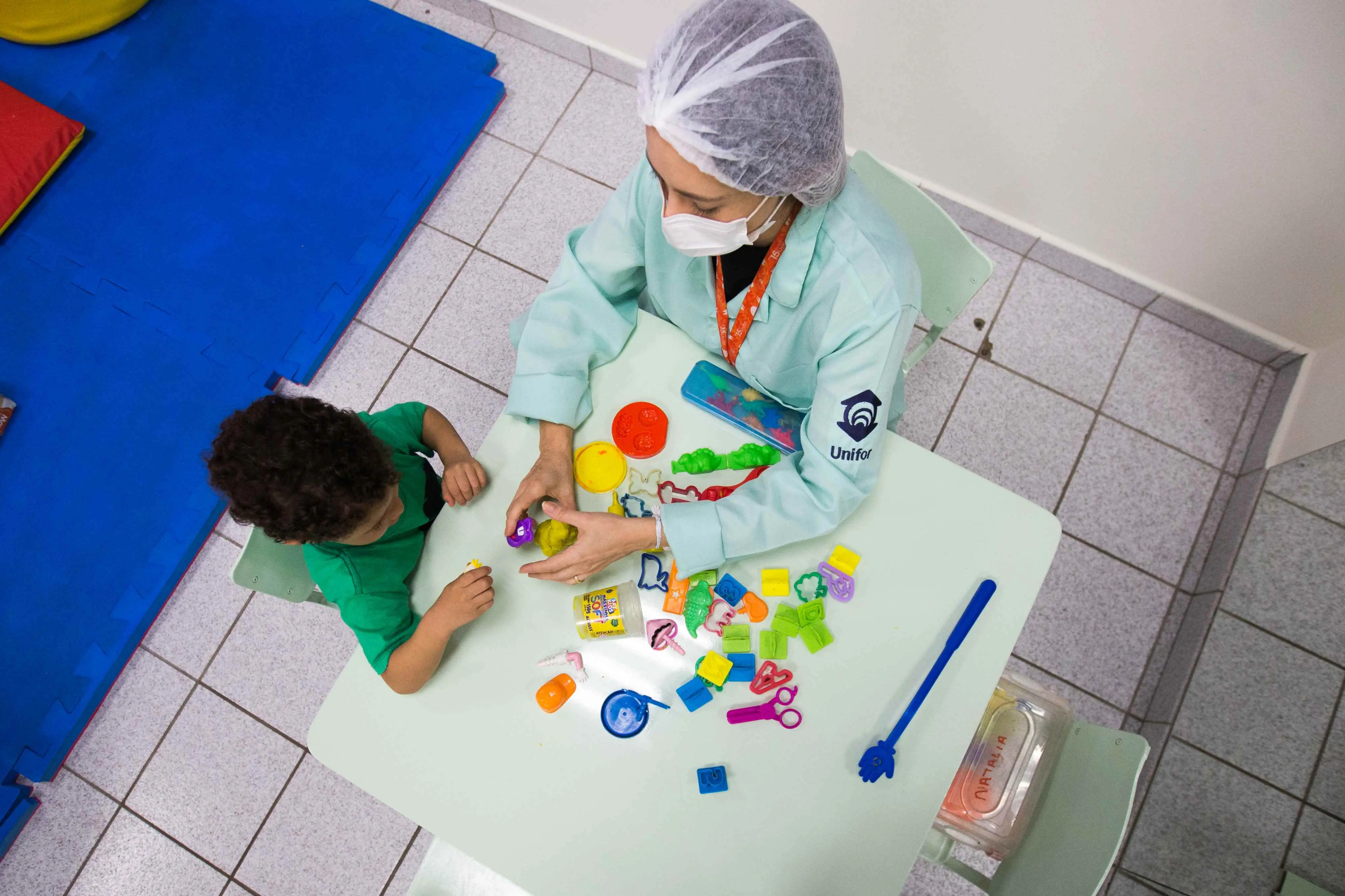 Vista aérea de uma profissional de saúde usando jaleco, máscara e touca, interagindo com uma criança em uma mesa branca. Ambos manuseiam massinha de modelar e moldes coloridos.