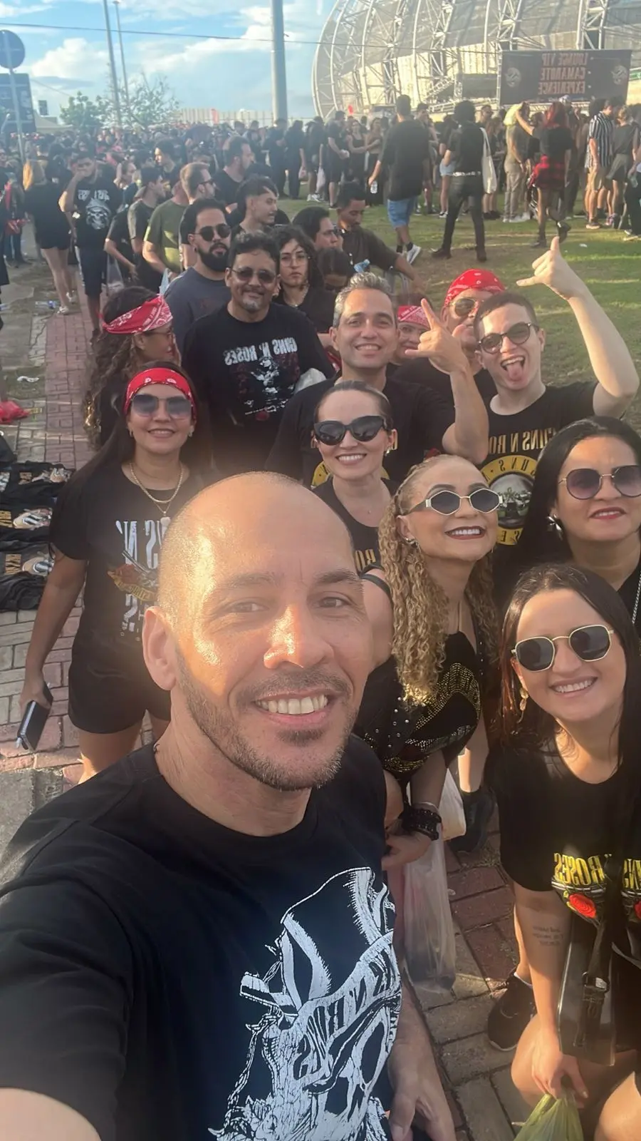 Selfie de um grupo de pessoas usando camisetas pretas em um evento ao ar livre, em fila, com grande público ao fundo e uma estrutura de palco visível ao longe.