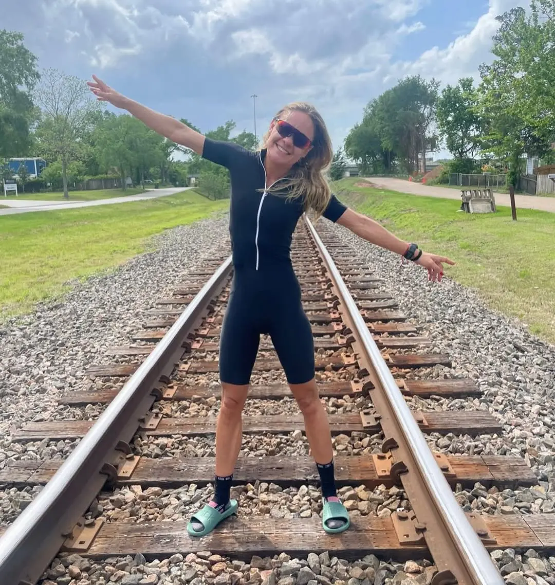 Mara, mulher magra, branca e loira, sorri para uma foto com os braços abertos.