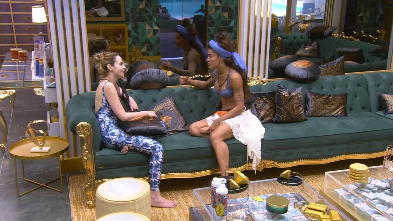 Duas pessoas sentadas em um sofá verde conversam em sala interna decorada, com almofadas estampadas, mesa de centro com objetos e espelho refletindo o ambiente ao fundo.