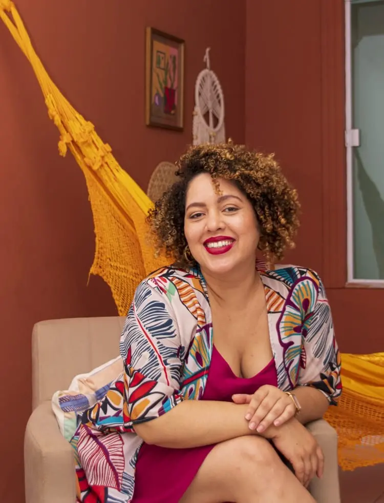 Na imagem, retrato de uma mulher de pele clara e sorriso aberto, sentada em uma poltrona bege. Ela tem cabelos cacheados curtos com luzes douradas e usa um batom vermelho vibrante. Veste um vestido magenta sob um quimono estampado com motivos de folhagens coloridas em tons de azul, laranja e amarelo. Suas mãos estão cruzadas sobre o joelho. O ambiente ao fundo tem uma parede cor de terracota, onde estão pendurados um quadro pequeno e um filtro dos sonhos branco. À esquerda, vê-se parte de uma rede de descanso amarela vibrante. A composição transmite uma sensação calorosa e acolhedora.