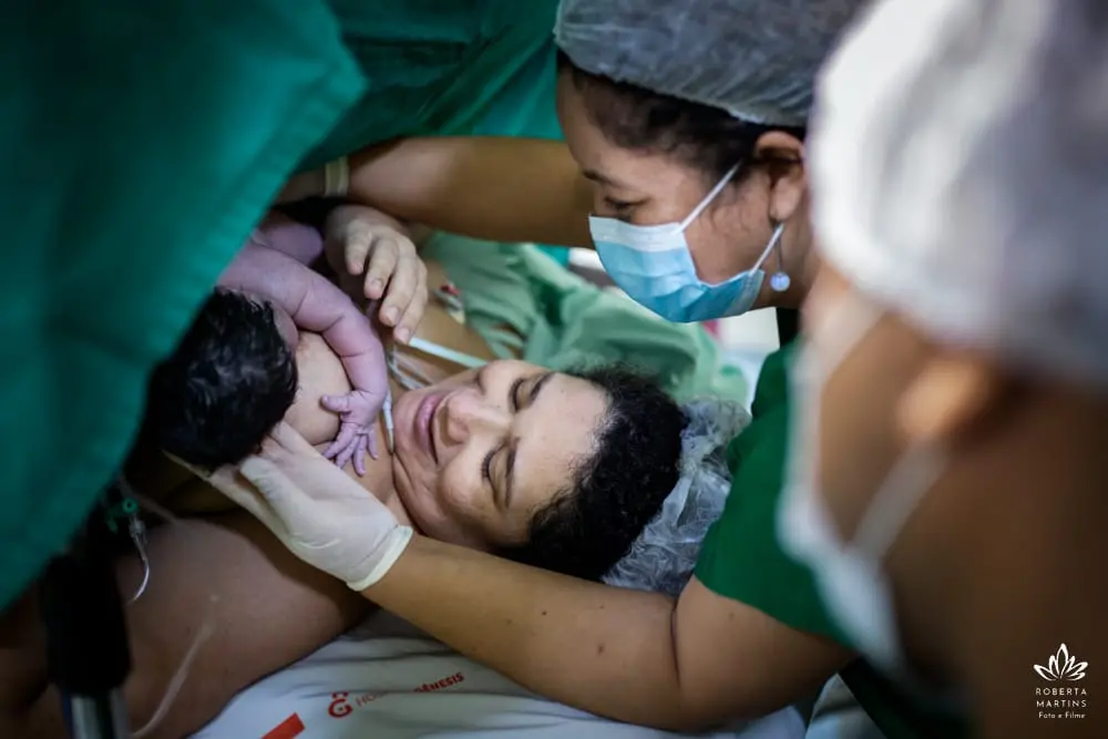 Na imagem, fotografia em ângulo superior de um momento de pós-parto imediato em um hospital. Uma mulher está deitada, com os olhos fechados e uma expressão de profundo alívio e serenidade, enquanto segura o seu bebê recém-nascido junto ao peito. O bebê, de cabelos escuros, está em contato pele a pele com a mãe. Ao lado, uma profissional de saúde (ou doula) de máscara azul e roupa verde observa a cena com carinho, amparando gentilmente o rosto da mãe com uma das mãos enluvadas. A cena é emoldurada por tecidos cirúrgicos verdes e outros profissionais em segundo plano, focando na conexão emocional entre mãe e filho. No canto inferior direito, há a logomarca 