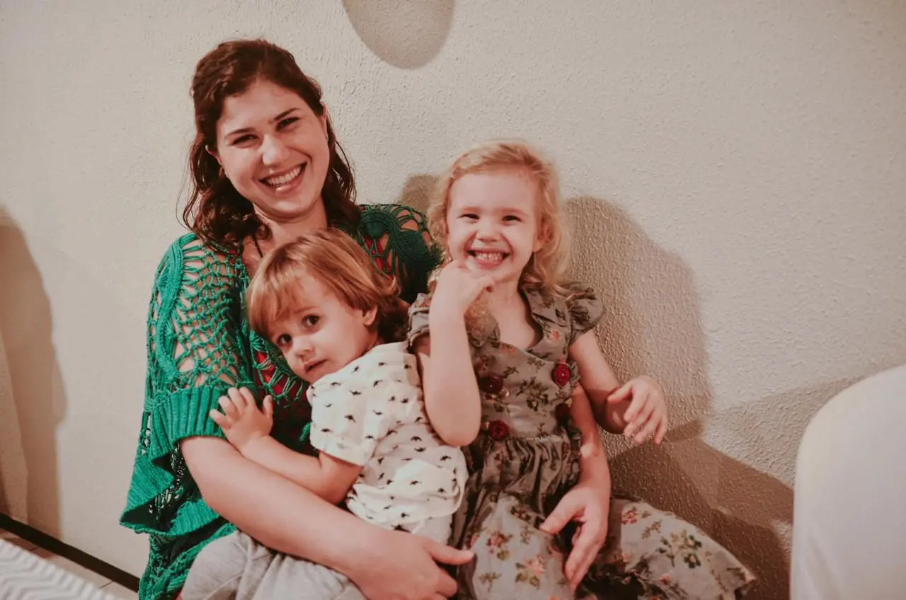 Na imagem, fotografia em plano médio de uma mulher jovem e sorridente sentada, abraçando duas crianças pequenas no colo. A mulher tem pele clara, cabelos castanhos ondulados e veste uma blusa de tricô verde com detalhes em crochê. À sua frente, no colo, um menino de cabelos castanhos curtos e camiseta estampada olha para a câmera. Ao lado dele, uma menina loira de cabelos cacheados e vestido floral cinza sorri abertamente, com a mão próxima ao rosto. Todos estão posicionados contra uma parede de cor neutra, em um ambiente interno com iluminação suave e quente, transmitindo um momento de carinho e alegria em família.