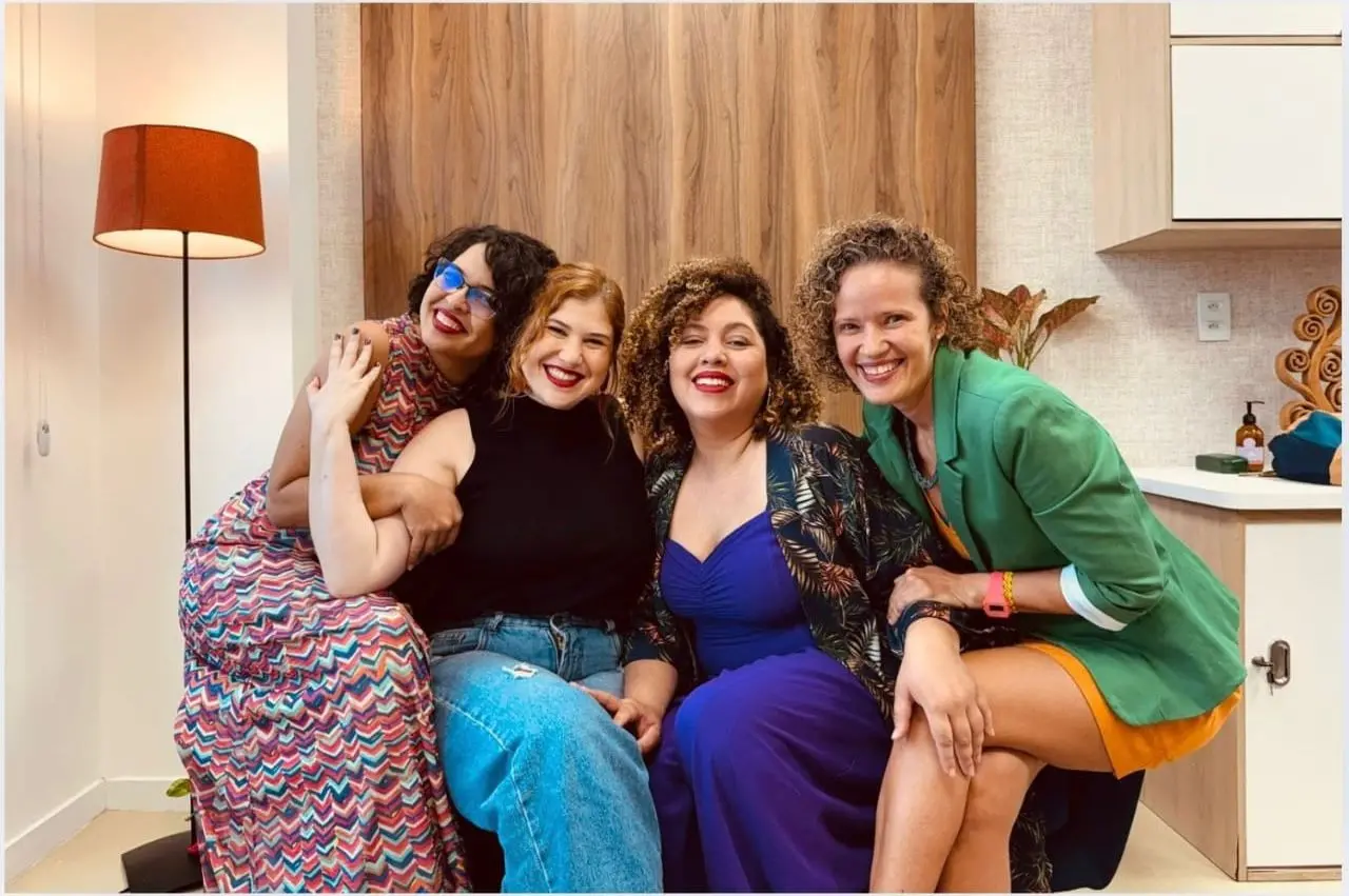 Na imagem, fotografia colorida de quatro mulheres sorridentes, posando juntas em um ambiente interno acolhedor. Elas estão sentadas próximas umas das outras, transmitindo união e amizade. Da esquerda para a direita: a primeira mulher tem cabelos curtos escuros, usa óculos de armação azul e um vestido longo com estampa geométrica colorida; a segunda tem cabelos ruivos, usa um top preto e calça jeans; a terceira tem cabelos cacheados volumosos, veste um macacão azul royal e um quimono estampado; a quarta mulher tem cabelos cacheados curtos e veste um blazer verde sobre uma roupa laranja. Ao fundo, há um painel de madeira, uma luminária de pé com cúpula laranja à esquerda e parte de um armário planejado à direita. A iluminação é clara e a atmosfera é de celebração e coletividade.