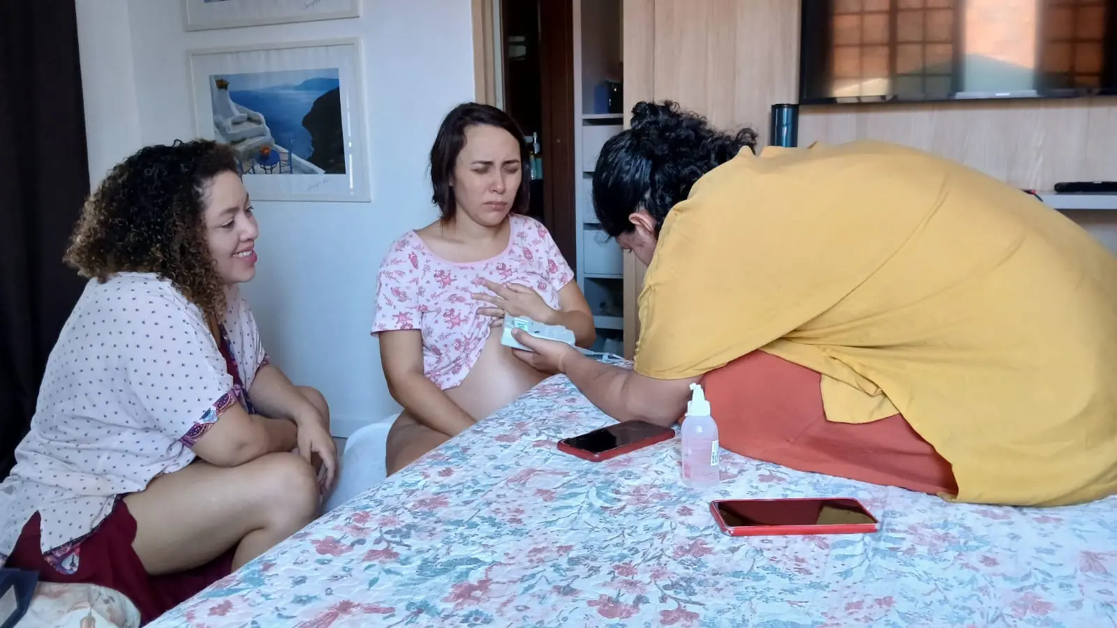 Na imagem, fotografia em plano médio de uma cena de assistência ao parto em um quarto. No centro, uma mulher grávida está sentada na cama, vestindo um pijama rosa estampado; ela tem os olhos fechados e uma expressão de concentração ou dor durante uma contração. À esquerda, uma mulher de cabelos cacheados (doula) observa a gestante com um sorriso encorajador e acolhedor. À direita, outra profissional de saúde, vestindo uma blusa amarela, está inclinada sobre a cama, utilizando um sonar Doppler para monitorar os batimentos cardíacos do bebê na barriga da gestante. Sobre a colcha floral da cama, veem-se dois celulares e um frasco de álcool em gel. Ao fundo, a decoração inclui um quadro com uma paisagem litorânea e uma televisão desligada.