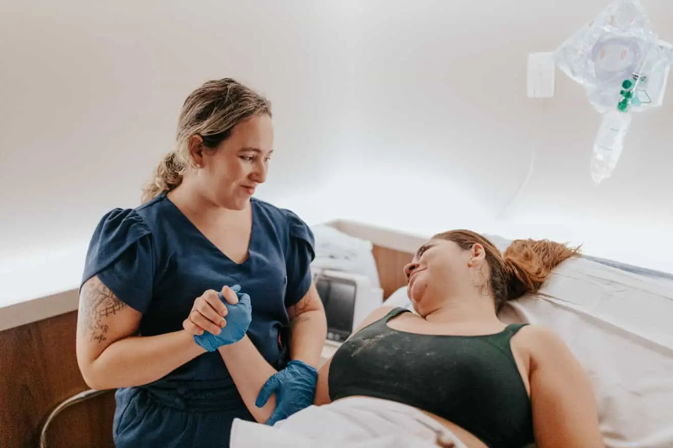 Na imagem, fotografia em plano médio mostrando um momento de cuidado e conexão em ambiente hospitalar. À esquerda, uma profissional de saúde de cabelos claros presos, usando um conjunto azul e luvas cirúrgicas azuis, segura a mão e o antebraço de uma mulher que está deitada na cama à direita. A mulher deitada, vestindo um top preto, olha para a profissional com uma expressão de confiança e serenidade. O ambiente é iluminado e sóbrio, com equipamentos médicos visíveis ao fundo, reforçando a atmosfera de assistência humanizada e acompanhamento durante o trabalho de parto.