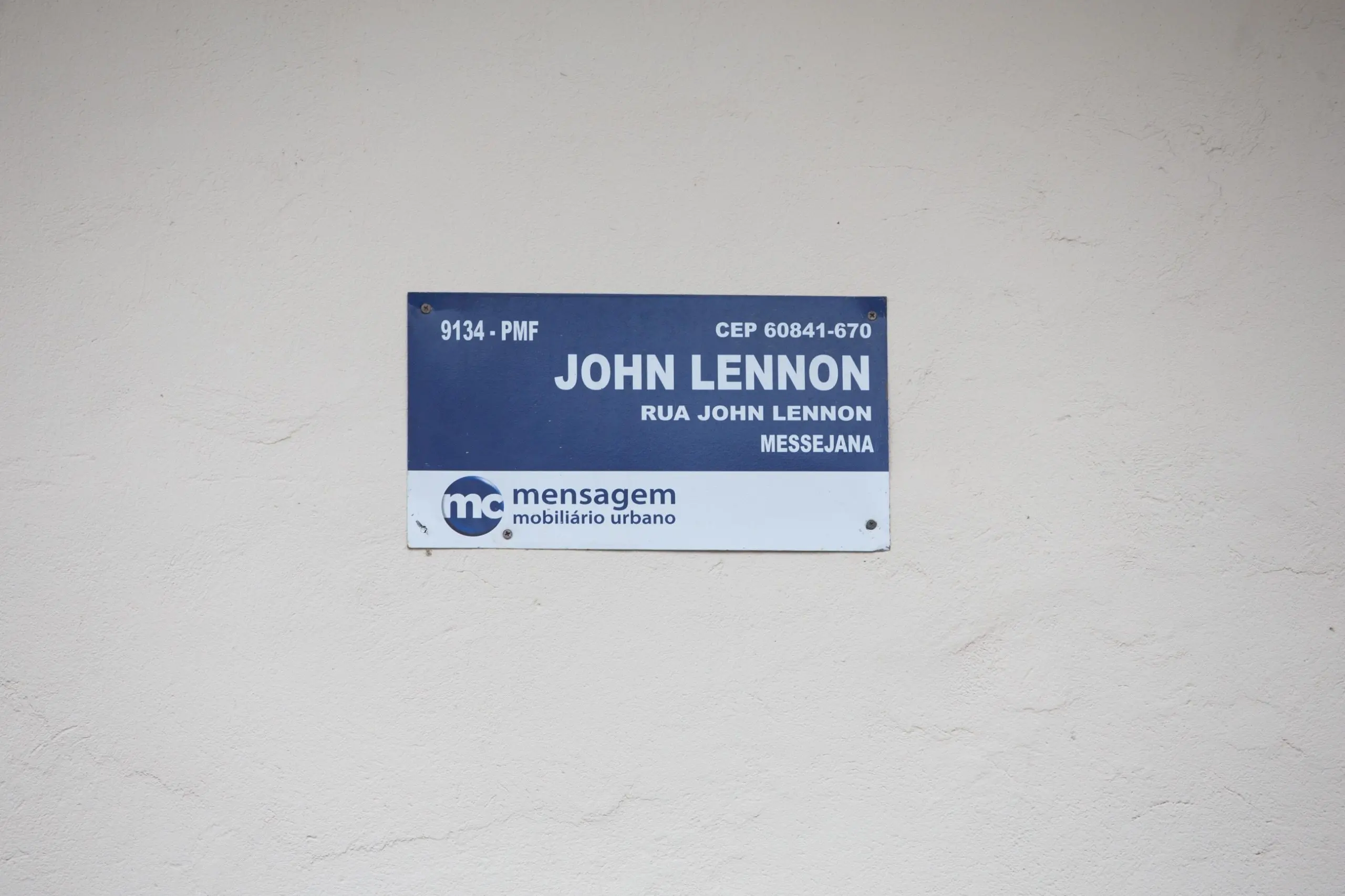 Placa azul da Rua John Lennon, no bairro Messejana, em Fortaleza, fixada em parede branca. O sinal exibe o nome do logradouro, o CEP e o logotipo da empresa de mobiliário urbano.