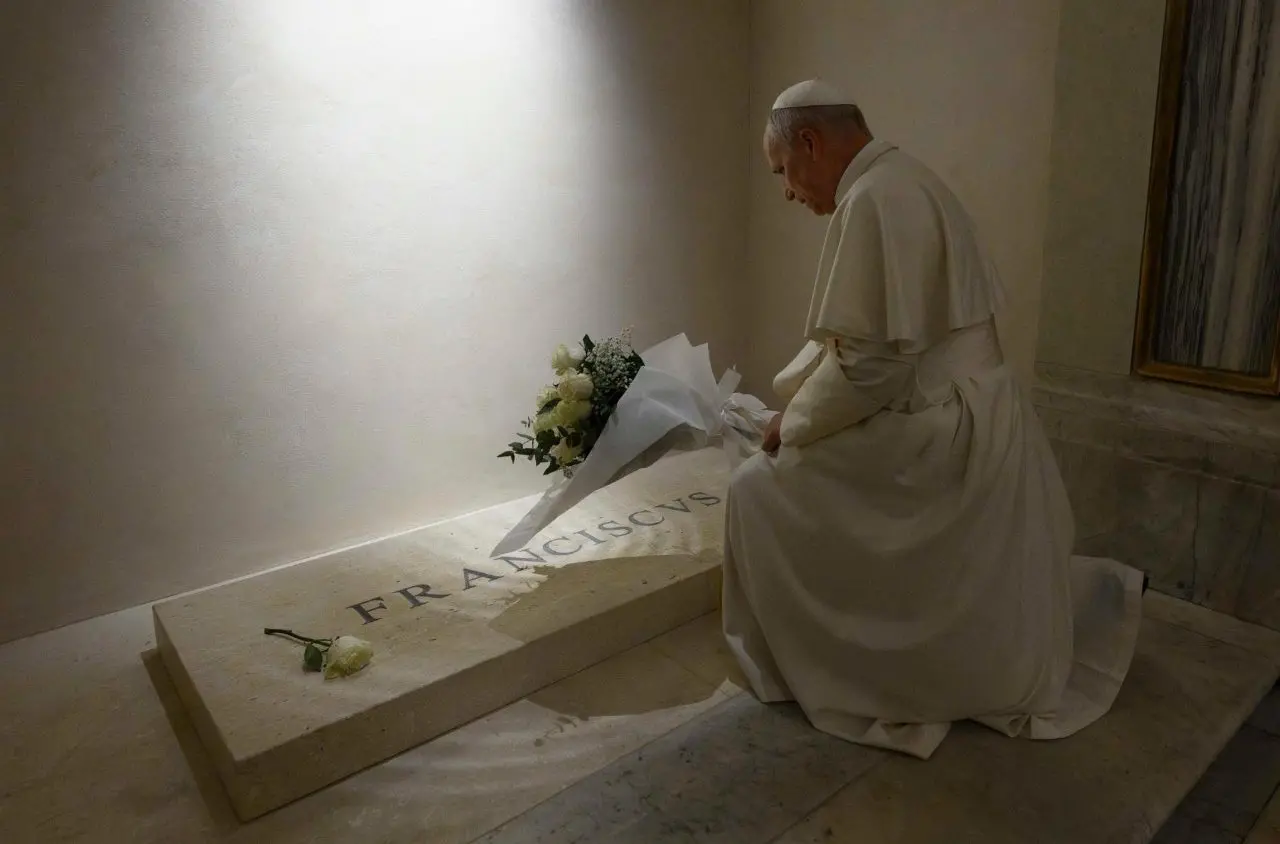 Papa Leão XIV ajoelhado em prece diante de um túmulo de mármore de papa Francisco. Ele segura flores brancas em um ambiente de iluminação suave e solene.