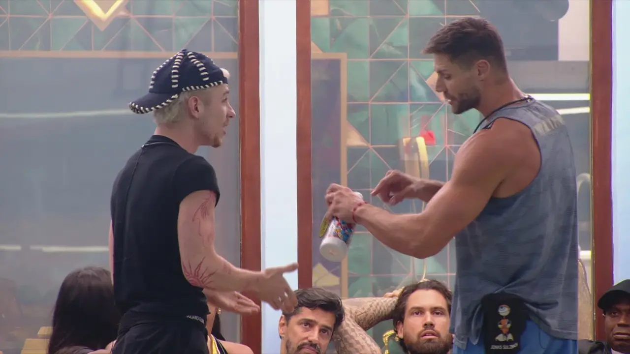 Duas pessoas em pé conversando frente a frente na área interna da casa de reality show, uma segurando uma garrafa; outras pessoas sentadas aparecem ao fundo.