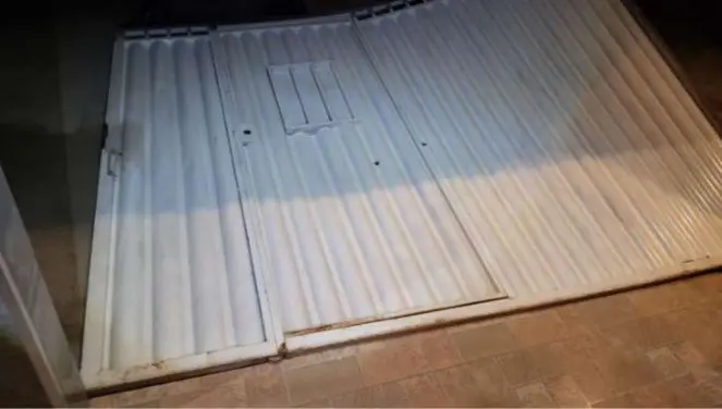 Imagem de um portão de garagem de metal branca, com uma entrada de acesso e fechadura, localizado na fachada de uma residência com piso de cerâmica.