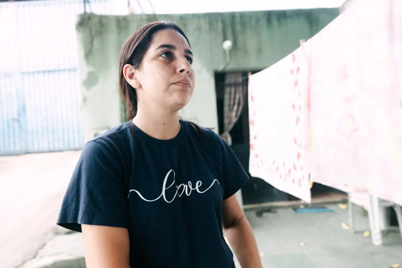 Mulher jovem branca olha para cima com expressão pensativa. Veste camiseta preta com a palavra Love em cursivo. Ao fundo, uma parede verde desgastada e roupas estendidas em um varal.