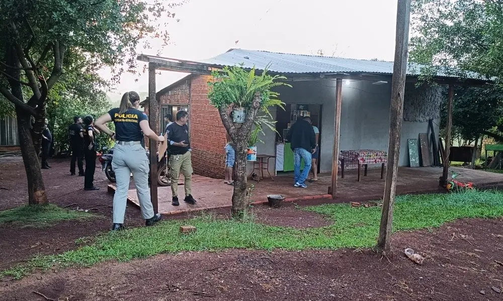 Policiais civis e agentes da Cooperação Internacional investigam uma residência rural de alvenaria com varanda simples, cercada por árvores e solo de terra vermelha. A cena registra a movimentação das autoridades na área externa e interna da casa durante o dia, em uma composição que destaca a perícia técnica no local.