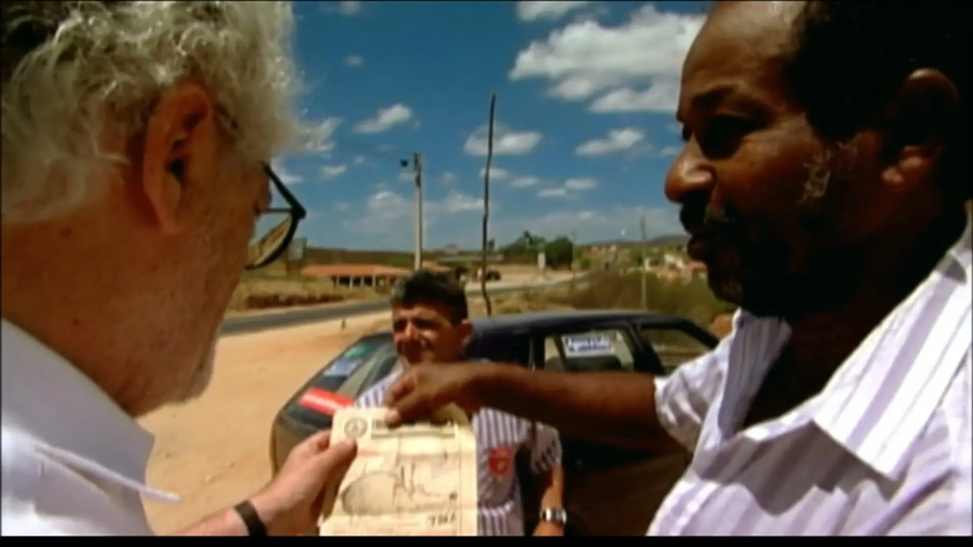 O cineasta Eduardo Coutinho, de perfil e cabelos brancos, observa um documento antigo apresentado por Zé Pretinho, ex-metalúrgico cearense, em uma estrada rural sob o céu ensolarado. A cena registra a troca de memórias entre os dois, com um carro estacionado e a paisagem árida ao fundo, destacando a importância do registro histórico pessoal.