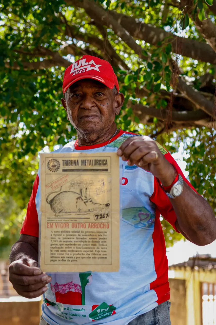 Zé Pretinho, um homem negro idoso, posa sob a copa de uma árvore, segurando com as duas mãos um exemplar preservado do jornal 'Tribuna Metalúrgica'. Ele veste um boné vermelho com a inscrição 'Lula 2022' e uma camiseta personalizada, mantendo um olhar sereno e direto para a câmera enquanto exibe o registro histórico.