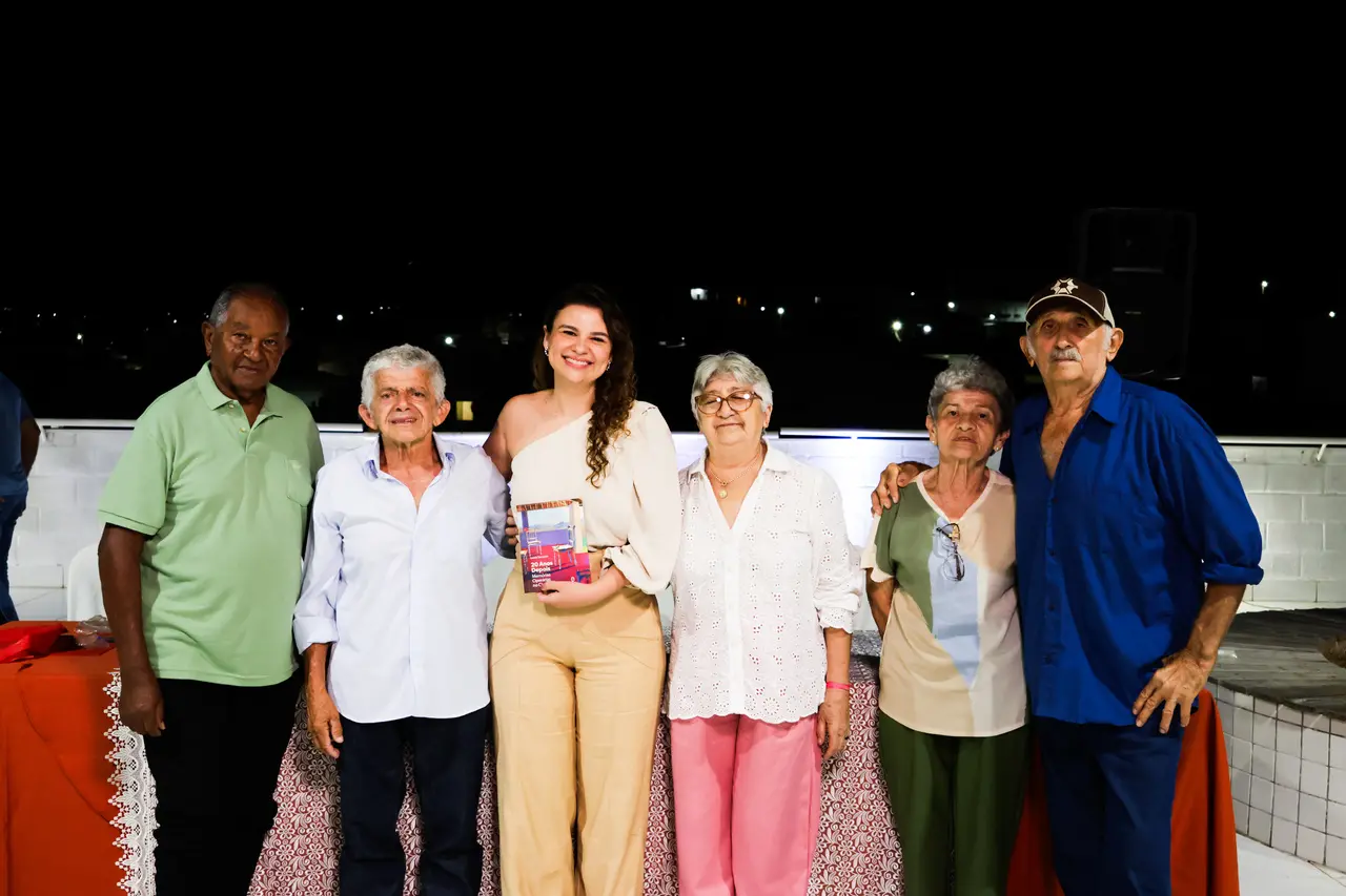 Uma mulher sorridente ao centro segura um livro e posa ao lado de cinco idosos em um ambiente externo à noite. O grupo está alinhado lado a lado à frente de uma mureta branca, com luzes urbanas distantes compondo o fundo sob o céu escuro.