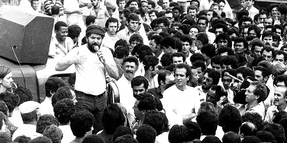 Lula discursa ao microfone em cima de um carro de som, cercado por uma multidão densa de trabalhadores durante uma assembleia histórica. A fotografia em preto e branco captura a energia do movimento sindical no ABC paulista, destacando o líder jovem de barba e boina em meio a um mar de rostos atentos.