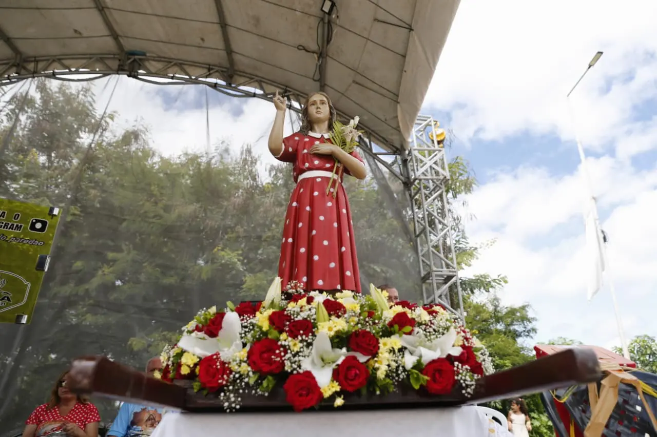 Imagem de uma celebração ao ar livre com uma estátua decorativa de uma mulher vestida de vermelho com bolinhas brancas, segurando um ramo de flores, rodeada por uma base de flores vermelhas e brancas. A celebração ocorre sob um palco com estrutura metálica e um céu parcialmente nublado.