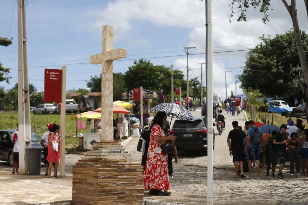 Cena em uma praça com uma cruz de madeira ao centro, pessoas com guarda-chuvas, carros estacionados e uma multidão participando de um evento religioso em um dia nublado.
