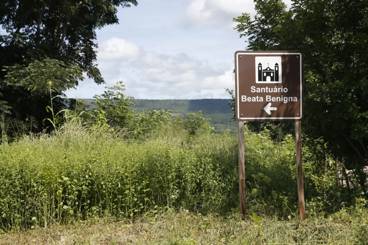 Sinalização indicando o caminho para o Santuário Beata Benigna em um cenário natural com vegetação e céu nublado ao fundo.