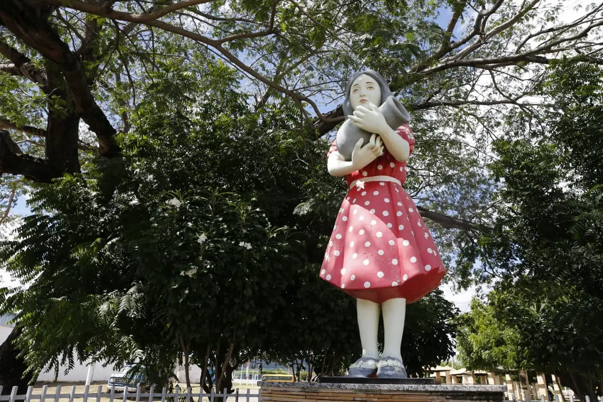 Estátua de uma menina de vestido rosa com poá branco segurando um gato sob um céu claro rodeada por árvores verdes. Ideal para buscas por esculturas ao ar livre.