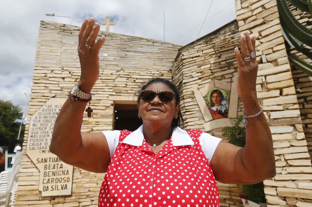 Mulher negra, vestida com blusa de bolinhas vermelha e branca, usando óculos escuros, fazendo um gesto com as mãos em frente a uma igreja ou capela, com uma imagem de uma santa na parede de fundo.