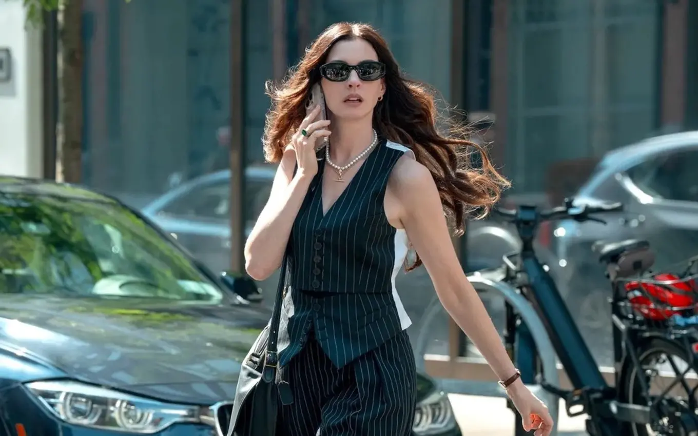 Pessoa andando em uma rua urbana, usando óculos escuros e roupa social sem mangas em tom escuro, com carros estacionados e bicicletas ao fundo, em ambiente externo durante o dia.