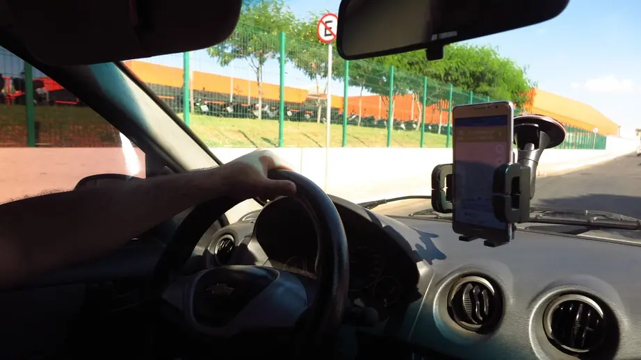 Pessoa dirigindo um carro com uma das mãos no volante enquanto o celular está preso a um suporte na frente do motorista.