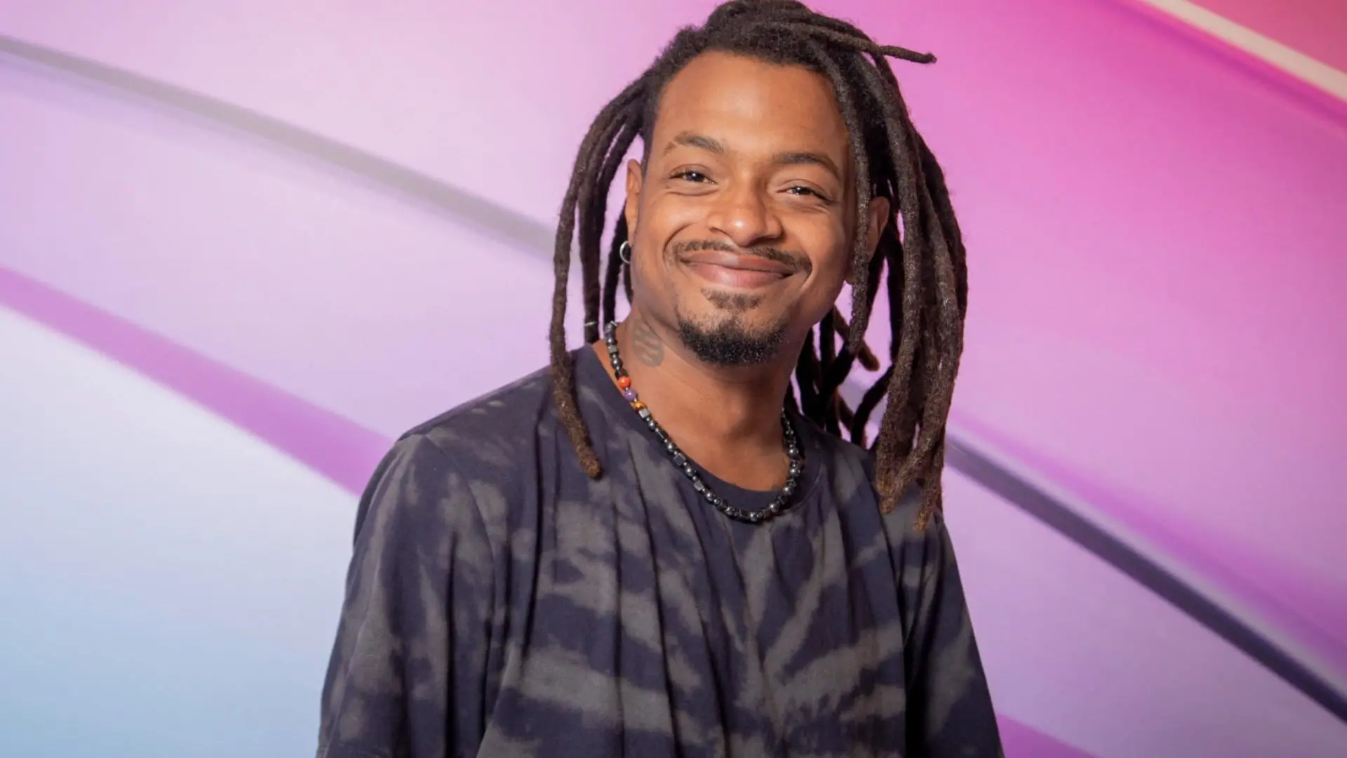 Homem com dreadlocks e sorriso, usando camiseta escura e colar, em fundo colorido com tons de rosa e roxo, transmitindo positividade.