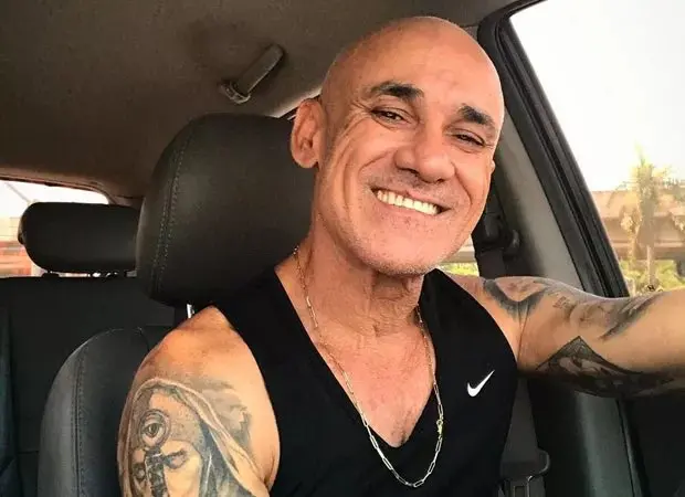 Homem sorrindo de tatuagens em um carro, usando uma regata preta e uma corrente, com expressão feliz e confiante.