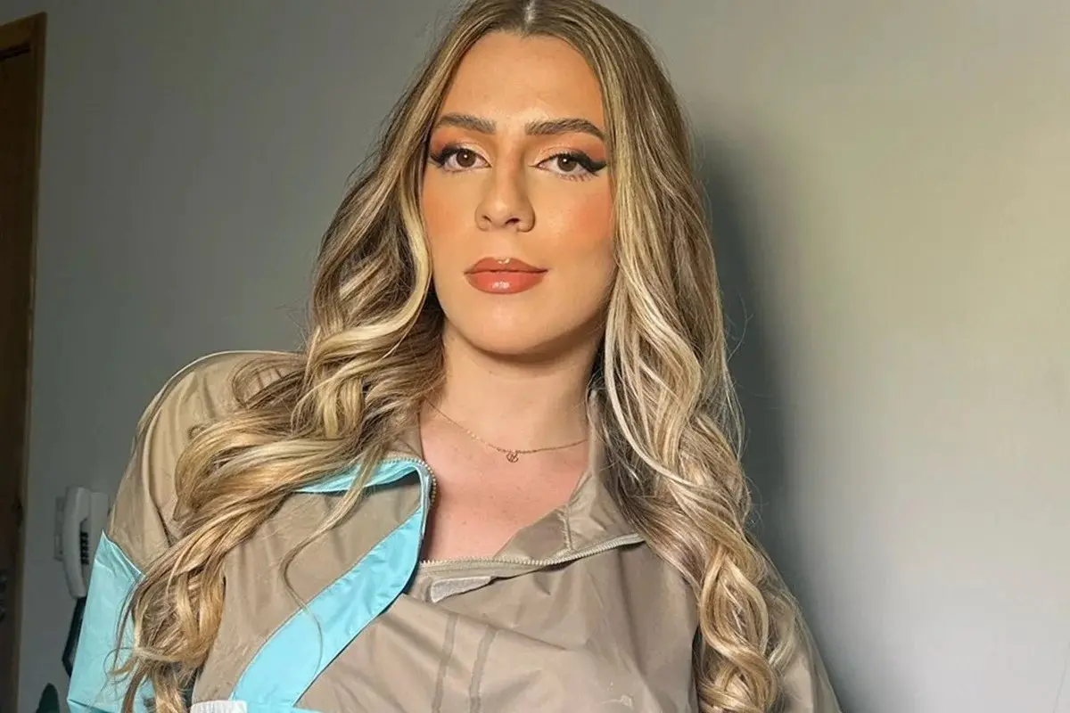 Mulher com cabelos cacheados e maquiagem natural, usando jaqueta bege com detalhes em azul, posando contra um fundo neutro. Fotografia de alta qualidade.